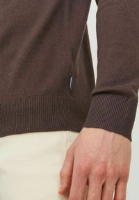 Maglione marrone a maglia con polsini e orlo a coste. Il tessuto appare morbido con una sottile texture. Piccolo logo del brand visibile sulla cucitura laterale.