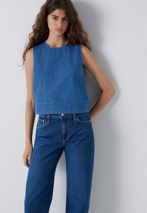 Woman with long wavy hair wearing a sleeveless blue denim top and high-waisted blue jeans standing against a plain white wall.