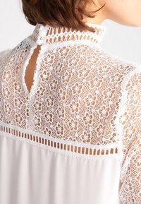 Blusa bianca con schiena in pizzo a motivi floreali, chiusura a bottoni al collo e parte anteriore in tessuto morbido. Contrasti di texture sottili.