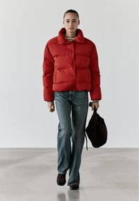 Rote Steppjacke mit hohem Kragen, horizontaler Steppung und Druckknopfverschluss, kombiniert mit blauen Jeans und einer dunkelbraunen Tasche. Glatte Textur.