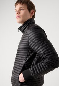 Melnā pūkainā jaka ar augstu apkakli, quilted dizainu un ribotām piedurknēm. Tajā ir sānu kabata un nemanāms logo uz piedurknes.