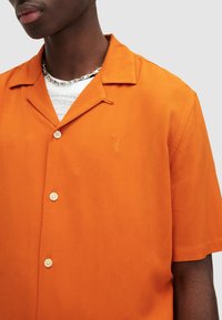 Camisa naranja de manga corta hecha de tejido ligero, con un cuello puntiagudo, dos botones y un pequeño diseño bordado en el pecho.
