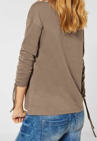 Brun långärmad skjorta med volanger vid ärmarna och knutdetaljer. Slät tyg och avslappnad passform, ihop med blå jeans som visar fickdetaljer.