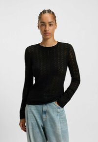 Pull noir à manches longues avec un motif zigzag texturé, silhouette ajustée et encolure ronde, associé à un jean clair ample.