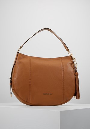 Brązowa skórzana torba na ramię Michael Kors z złotymi okuciami i detalem w postaci frędzli, prezentowana na białym pedestale na szarym tle.