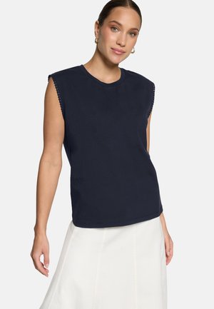 Kobieta o jasnej karnacji i zielonych oczach, nosząca złote kolczyki koła, granatowy bezrękawnik z ozdobnymi krawędziami oraz białą spódnicę.