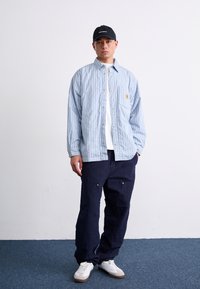 Chemise à rayures bleu clair et blanche, pantalon cargo marine, baskets blanches et une casquette noire avec le logo de la marque. Fond uni.