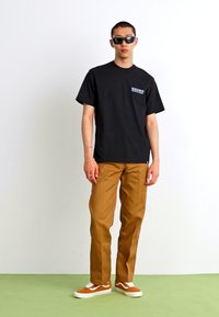 Svart bomull t-skjorte med lite logo, tan chinos, og brune stripete sneakers, stående mot en hvit bakgrunn med et grønt gulv.
