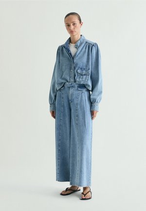 Vrouw draagt lichtblauwe spijkerjas en wijde jeans, gecombineerd met zwarte sandalen met kralen, staand tegen een eenvoudige witte achtergrond.