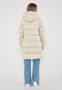 Giacca imbottita beige con cappuccio, caratterizzata da un design trapuntato e un taglio a lunghezza intermedia. Abbinata a jeans larghi blu e scarpe da ginnastica bianche.