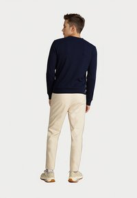 Férfi sötétkék pulóverben, bézs nadrágban és fehér sportcipőben áll, háttal egy sima világos háttérnek.