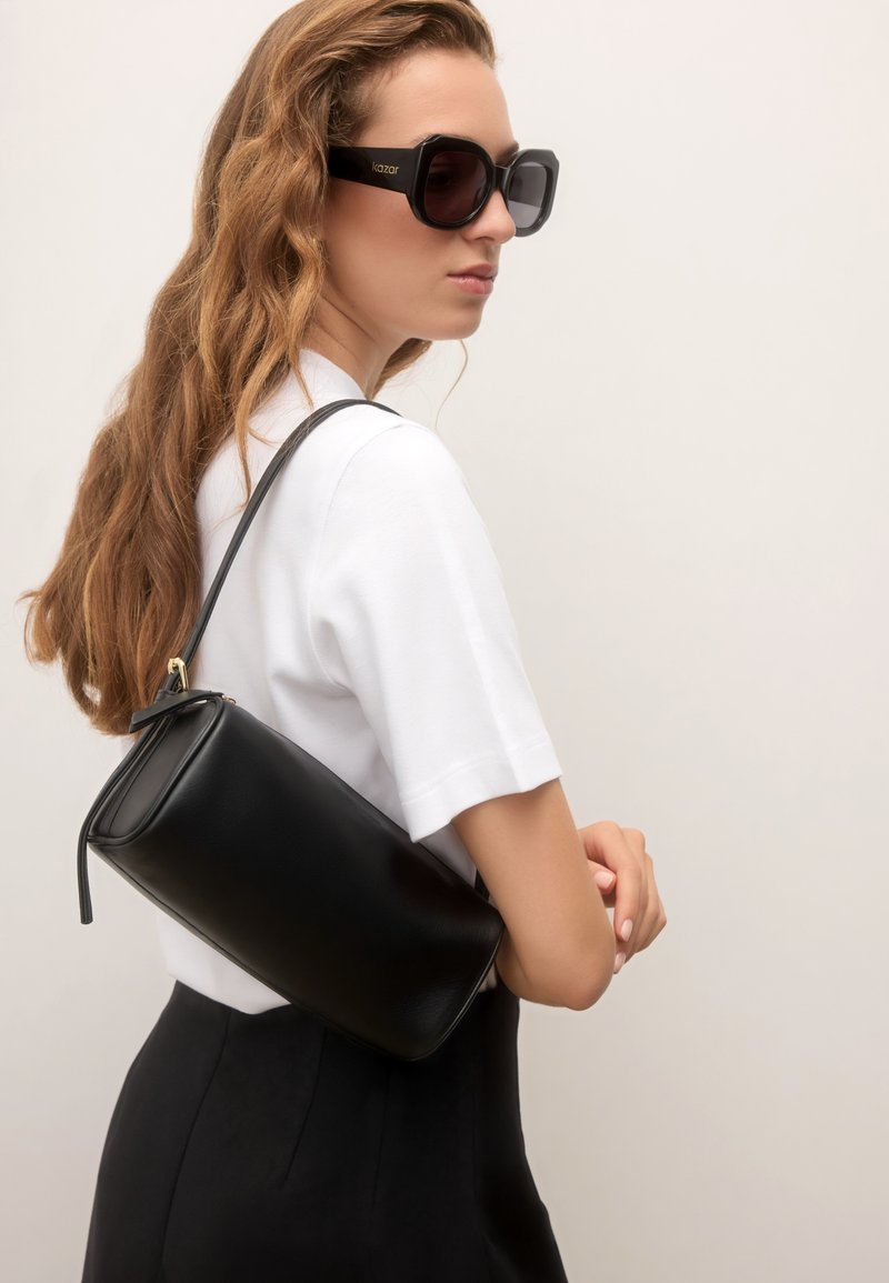 Femme aux longs cheveux portant de grandes lunettes de soleil noires, un haut blanc à manches courtes, un pantalon noir et un sac à bandoulière noir.