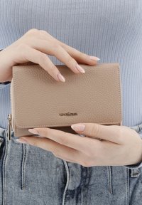 Béžová texturovaná kožená peněženka se zipem, logo na přední straně, obdélníkový tvar, držená oběma rukama proti světle modrému topu.