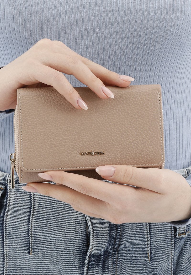 Béžová texturovaná kožená peněženka se zipem, logo na přední straně, obdélníkový tvar, držená oběma rukama proti světle modrému topu.