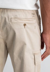 Beige broek met een achterzak met knoop, een zijvak en een klein gestikt logo bij de zak, gedragen door iemand in een wit shirt.