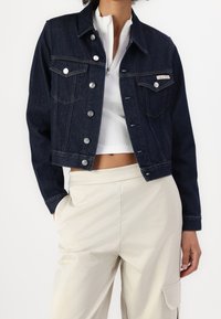 Tmavě modrá cropovaná džínová bunda s kovovými knoflíky, kapsami na prsou a kontrastním prošíváním, doplněná bílým žebrovaným topem a béžovými kalhotami.