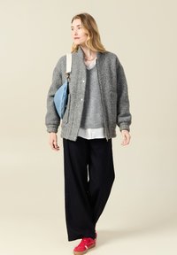 Graue Wollbomberjacke mit elastischen Bündchen, grauem Pullover, weißem Hemd, schwarzen weit geschnittenen Hosen und roten Sneakers. Blauer Beutel über der Schulter.