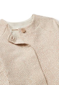 Krémová dětská kombinéza s malými hnědými puntíky, kulatým výstřihem a jedním hnědým knoflíkem nahoře.
