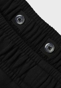 Juodas elastingas juosmuo su susiraukšlėjusia faktūra, kuriame yra du skaidrūs mygtukai prisiūti prie juodo audinio lopinėlio. Matomi lygaus paviršiaus detalės ir siuvinėjimas.