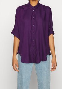 Lila knapp-up skjorta med krage, avslappnad passform, korta ärmar och slät textur, tillsammans med ljusblå jeans.