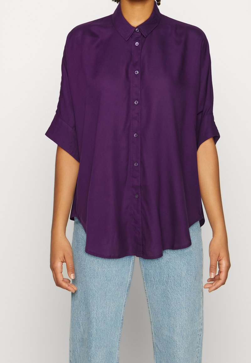 Lila knapp-up skjorta med krage, avslappnad passform, korta ärmar och slät textur, tillsammans med ljusblå jeans.