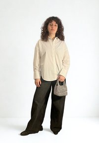 Biała koszula na guziki, czarne spodnie o szerokich nogawkach, teksturowana czarno-biała torba. Modelka ma kręcone włosy, stoi na jednolitym tle.