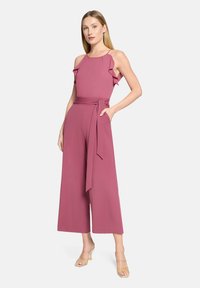 Ärmelloser Overall mit Rüschenbesatz in zartem Rosa, ausgestattet mit einem gebundenen Taillengürtel, weitem Bein und seitlichen Taschen. Glatter Stoff mit leichter Textur.