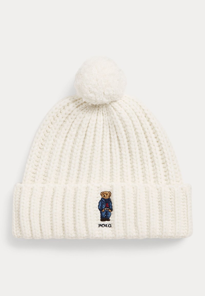 Bonnet blanc tricoté avec un pompon sur le dessus et un petit ours brodé portant une veste bleue sur le revers avant.