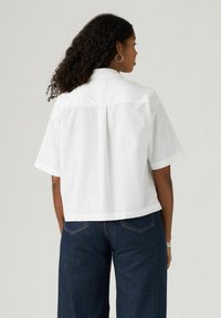 White short-sleeve shirt with a back yoke and pleat detail, designed with a relaxed fit. Worn with dark blue wide-leg jeans.