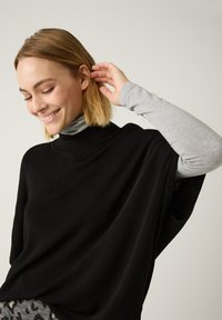Schwarzer, oversized Pullover mit hohem Kragen, über einem hellgrauen Langarmshirt getragen. Weiche Stricktextur, lockere Passform, minimalistisches Design.