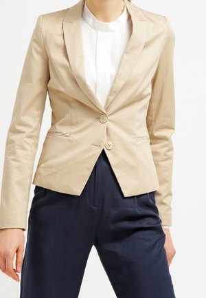 Veste ajustée beige avec deux boutons, revers crantés et poches latérales, associée à un pantalon marine foncé. Tissu en mélange de coton doux.