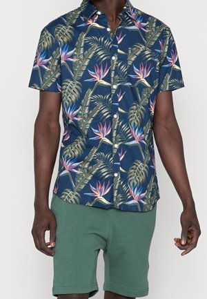 Homme portant une chemise boutonnée à manches courtes bleu marine avec des feuilles et des fleurs tropicales, associée à un short vert.