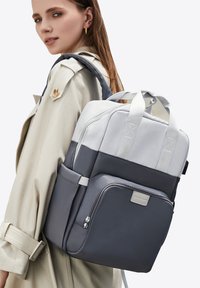 Sac à dos gris et gris clair avec deux poignées supérieures, poche zippée à l'avant et bretelles rembourrées, présentant une texture lisse et mate.