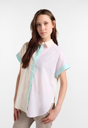 Junge Frau trägt ein kurzärmeliges Hemd mit Pastellstreifen in Creme, Pink und Mintgrün, kombiniert mit hellbeigen Hosen.