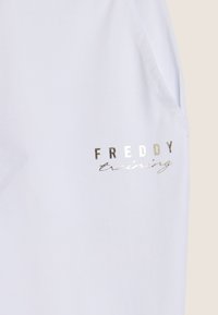 Top atlético blanco con un estampado metálico plateado de "FREDDY training" en el lado izquierdo, con una textura suave y mangas cortas.