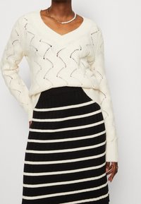 Pull en maille crème avec col en V et motif ondulé, associé à une jupe côtelée rayée noire et blanche. Tissus texturés et design ajusté.
