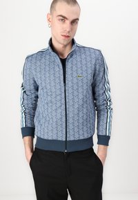 Jachetă bomber bleumarin matlasată cu model geometric, cu mâneci cu dungi contrastante și fermoar frontal. Prezintă un logo verde pe piept.