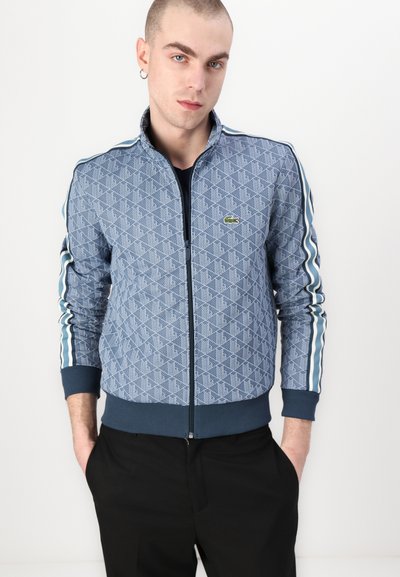 Veste bomber bleue matelassée avec un motif géométrique, des manches à rayures contrastantes et une fermeture éclair à l'avant. Présente un logo vert sur la poitrine.