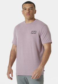 Camiseta corta de manga corta de color púrpura claro, hecha de una tela suave, con un pequeño logo negro en el área del pecho izquierdo y un escote redondo.