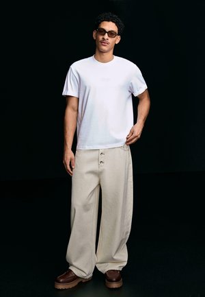 Moški, oblečen v belo majico, ohlapne bež hlače z gumbi, rjavi čevlji in sončna očala, stoji ob črni ozadju in si z eno roko prilagaja hlače.