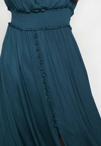 Tyrkis klänning med smockad överdel och rufflade kanter. Har en knäppt slitsöppning längs sidan och ett flödande tyg med mjuk textur.