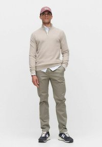 Ljusbeige tröja med kvartdragkedja, bärs över en vit skjorta. Olivgröna byxor och mörka sneakers med vita detaljer fulländar outfiten.