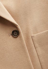 Khakifarbener Wollmantel mit strukturiertem Oberflächenmaterial, einem einzelnen dunklen Knopf und einer seitlichen aufgesetzten Tasche mit sauberem Stichdetail.