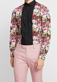 Chemise à manches longues à motif floral avec un panneau avant noir ; associée à un pantalon rose clair. Présente des textures variées et des couleurs vives.