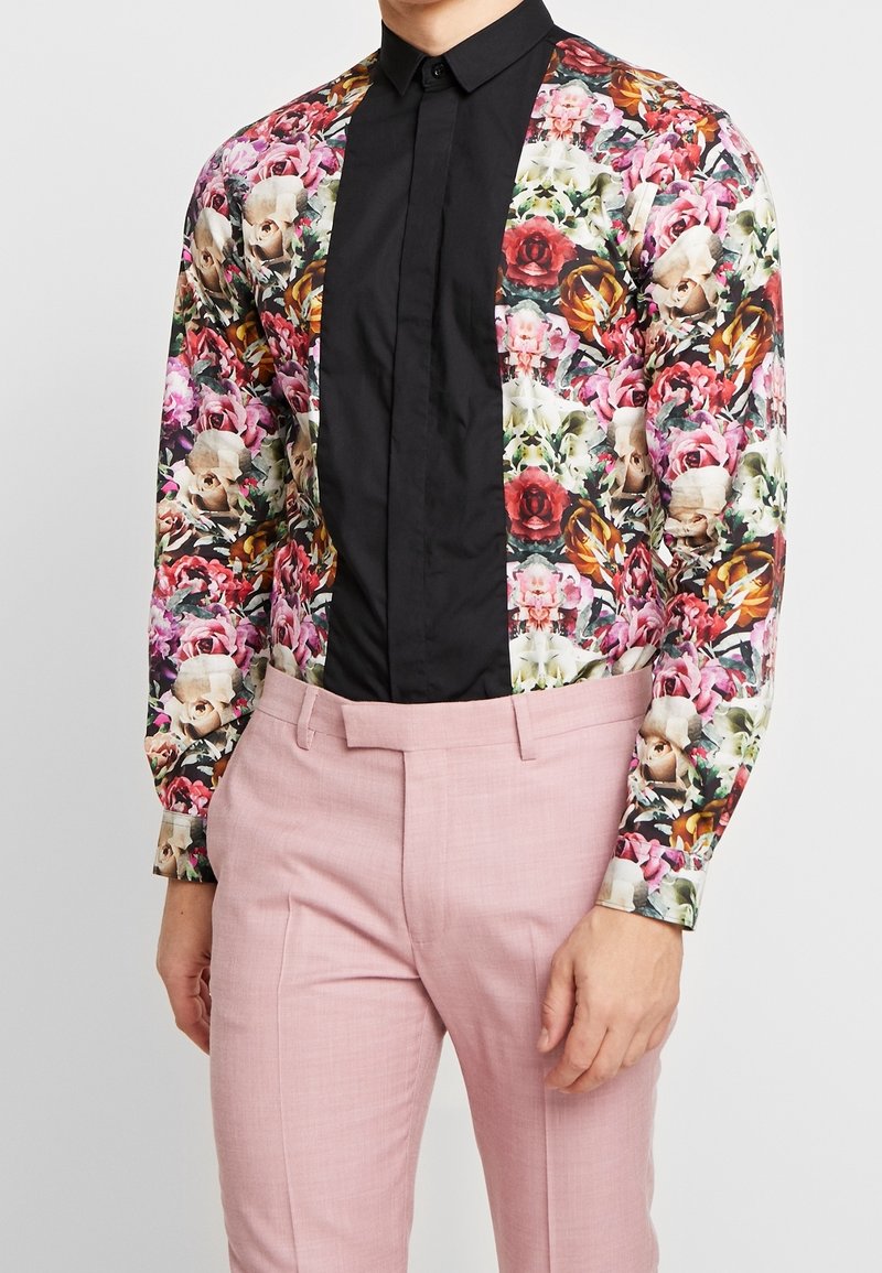 Chemise à manches longues à motif floral avec un panneau avant noir ; associée à un pantalon rose clair. Présente des textures variées et des couleurs vives.
