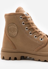 Botte cheville en toile marron avec un laçage, semelle en caoutchouc et surface texturée. Présente un patch logo circulaire sur le côté.