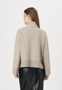 Beige genser med høy hals, løs passform og ribbestrikket kant. Glatt tekstur, designet med senket skuldre. Kombinert med et svart skjørt.