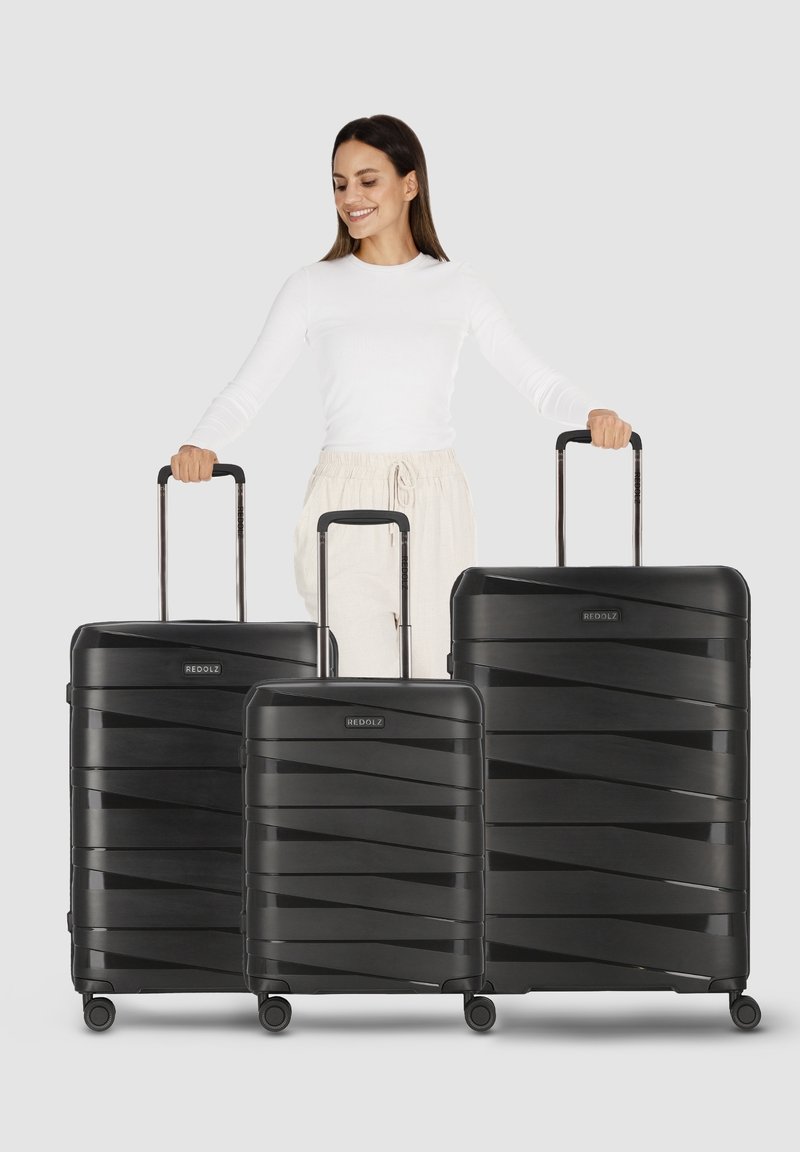 Femme en haut blanc et pantalon beige tenant les poignées de trois valises noires à roulettes de tailles variées devant un fond uni.