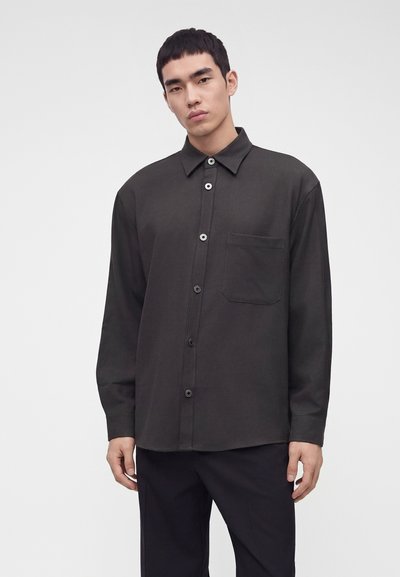 Jeune homme portant une chemise gris foncé à boutons avec une poche sur la poitrine, debout devant un fond clair uni.