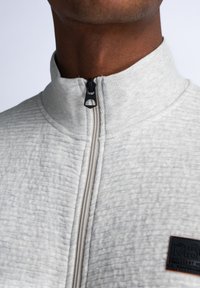 Ljusgrå zip-up jacka med strukturerad tyg, högkrage och kontrasterande svart dragkedja. Har en liten logotyp på framsidan.